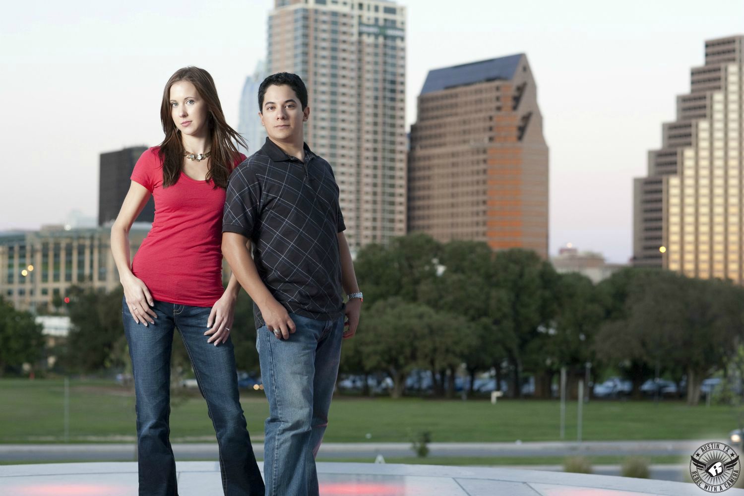Red headed, blue eyed girl wears a red scoop neck shirt with blue jeans and a necklace standing next to a brown haired Latino guy wearing a grey crosshatch and blue jeans at the Long Center with the city skyline of Austin in this impressive engagement session in Austin, Texas.  